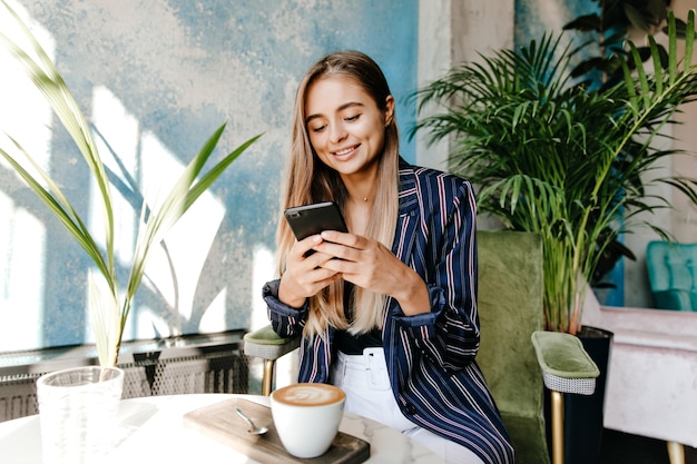 Messaggio di sms di donna bianca sbalorditivo mentre beve il cappuccino. amabile ragazza europea in giacca che riposa nella caffetteria.