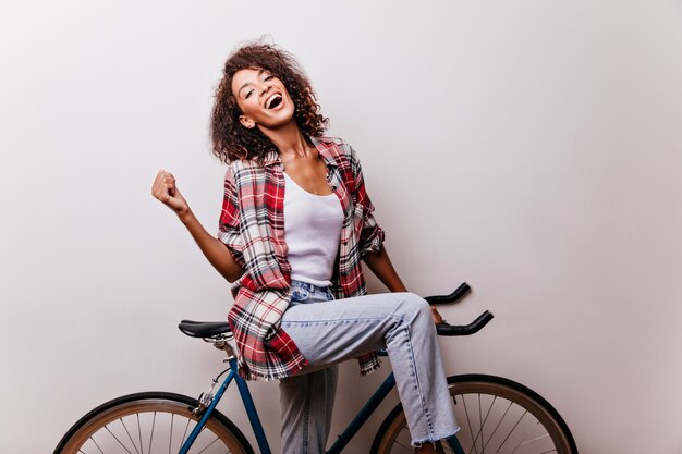 Потрясающая женщина в синих джинсах и красной рубашке позирует на велосипеде. Крытый снимок смеющейся мечтательной девушки, весело проводящей время.