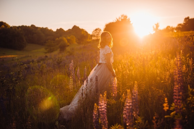 明るいドレスで見事な女性が夕日の光の中をフィールドを横切って歩く
