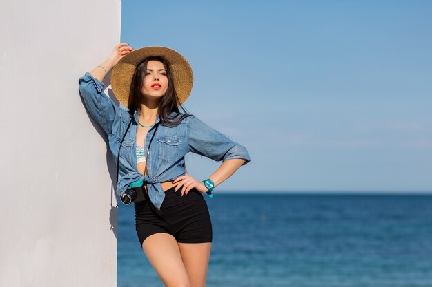 Stunning woman with figure in shorts and straw hat posing on the beach.