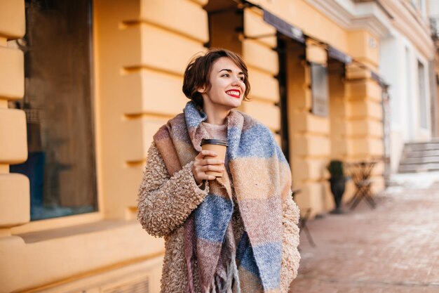 コーヒーを飲む長いスカーフを持つ見事な女性寒い日を楽しんでいるスタイリッシュな女の子の屋外ショット