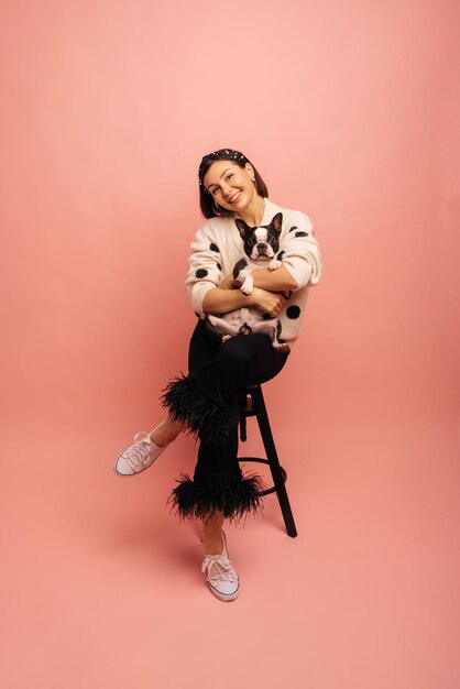 Stunning young caucasian brunette woman holding french bulldog while sitting on pink background Showing affection concept