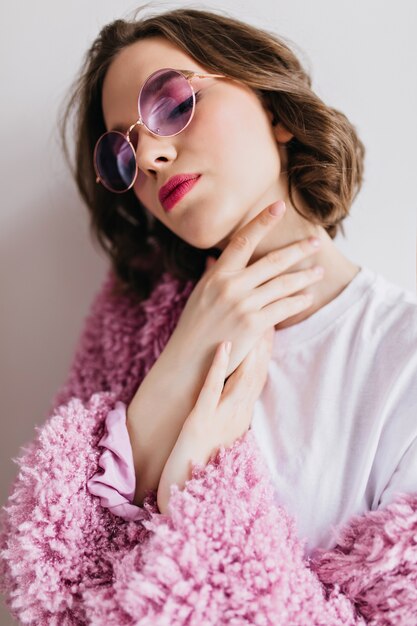 눈을 가진 포즈 보라색 선글라스에 멋진 젊은 아가씨는 흰 벽에 마감했다. 분홍색 모피 코트에 사랑스러운 곱슬 소녀의 근접 사진.