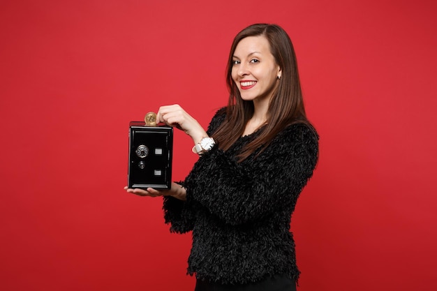 Stunning young woman in fur sweater holding metal bank safe for money accumulation, bitcoin, future currency isolated on red background. people sincere emotions, lifestyle concept. mock up copy space.