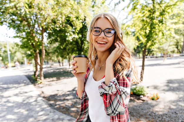 自然に一杯のコーヒーを保持しているメガネの見事な若い女性。夏の日に公園を歩いて笑顔のブロンドの女の子。