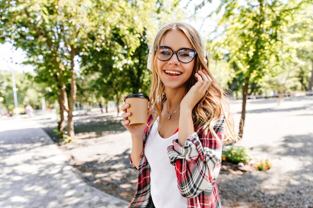 Stunning young woman in glasses holding cup of coffee on nature. Smiling blonde girl walking around park in summer day.