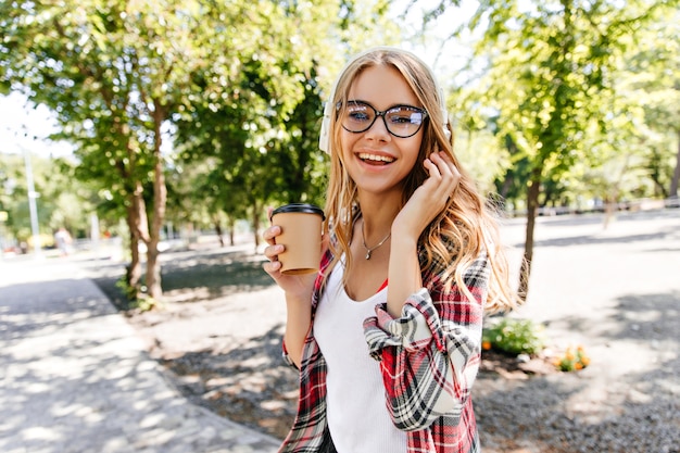 Потрясающая молодая женщина в очках, держа чашку кофе на природе. Улыбающаяся блондинка гуляет по парку в летний день.