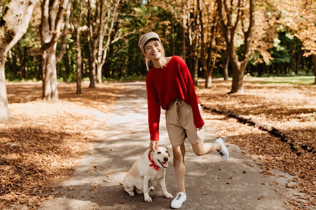 彼女の犬と一緒に美しい秋の公園で楽しんでいるトレンディな赤いセーターとベージュのショートパンツで見事な若い女性。