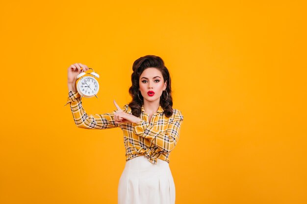 Stunning young woman with bright makeup posing with clock. Studio shot of appealing pinup girl isolated on yellow background.