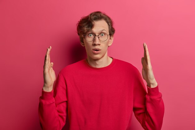 Stupefied amazed man youngster shapes huge object, makes something big, gasps from wonder, has surprised expression, measures and explains size, dressed casually, poses against pink wall