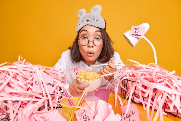 Stupefied Asian woman in domestic wear has breakfast at workplace eats cornflakes cannot believe her eyes sits at desktop with cut paper around isolated over yellow wall works from home