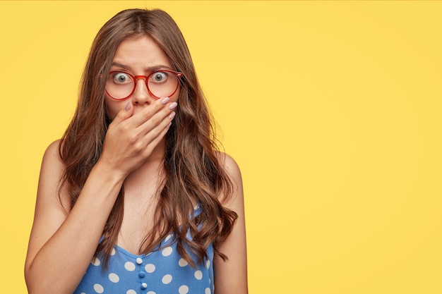 Stupefied attractive European woman covers mouth with palm, has frightened expression as notices something terrible