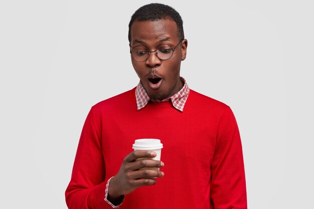 Ошеломленный темнокожий молодой человек делает перерыв на кофе, слышит потрясающие новости от собеседника, держит одноразовую чашку, затаив дыхание, носит красный свитер