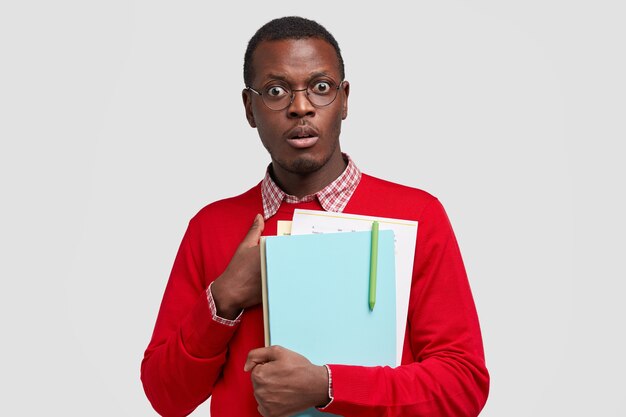 Stupefied dark skinned erudite being shocked to be asked unexpected question, holds textbooks and papers with pen