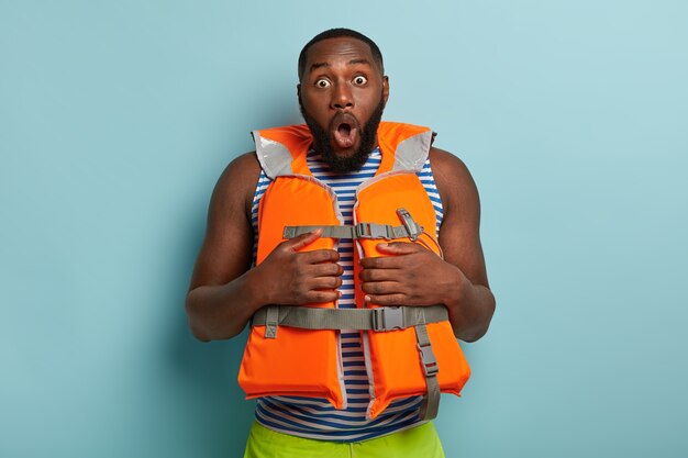 Stupefied dark skinned man dressed in life jacket, shocked to have emergency situation, has thick beard