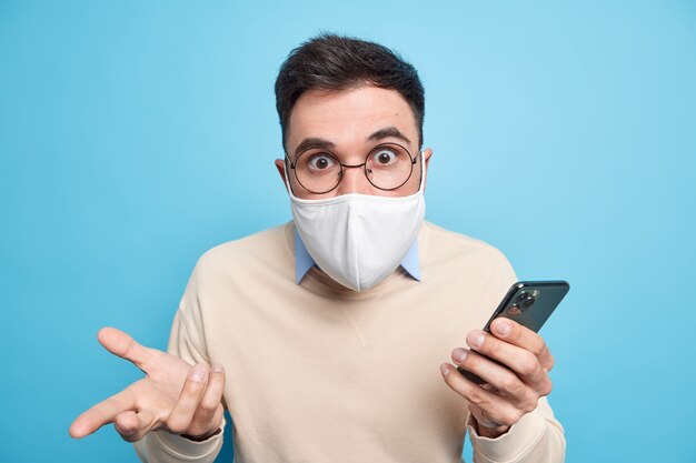 Stupefied embarrassed adult man stares shocked, wears mask to prevent getting infected by coronavirus stays safe during home quarantine uses mobile phone 