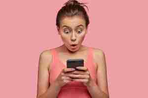 Free photo stupefied emotional woman stares with bugged eyes at smart phone, feels shocked as reads news at web page, connected to wireless internet, has break after training with coach in gym, wears pink top