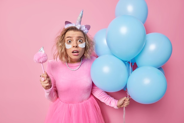 Stupefied frightened young woman stares at camera holds breath wears unicorn headband festive dress holds bunch of inflated blue balloons applies beauty patches for skin treatment being on party