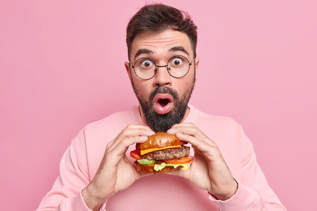 맛있는 식욕을 돋우는 패스트 푸드와 함께 포즈를 취하는 Stupefied 잘 생긴 남자는 맛있는 햄버거를 들고 둥근 안경을 쓰고 캐주얼 점퍼는 다이어트를 유지하지 않습니다.