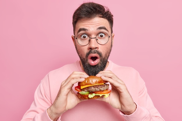 Ошеломленный красавец позирует с восхитительным аппетитным фастфудом, держит вкусный гамбургер, носит круглые очки, повседневный джемпер, не придерживается диеты, любит читмил, одетый небрежно