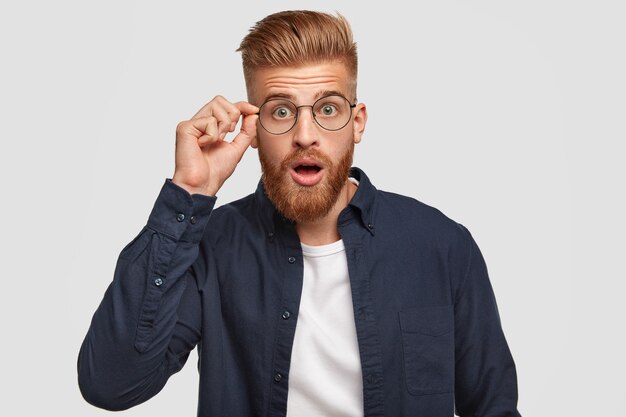 Stupefied hipster guy with shocked expression, hears something unbelievable, keeps hand on rim of round glasses, opens mouth from surprisement, dressed in stylish shirt, isolated on white wall