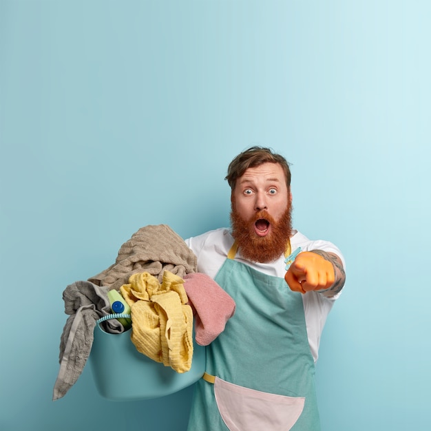 Free photo stupefied housekeeper points surprisingly at camera, wears blue apron, holds basin of linen, cannot believe eyes