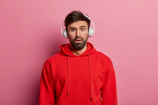 Stupefied male meloman stares surprisingly , listens audio via headphones, dressed in red sweatshirt, hears astonishing news, poses over rosy wall. People, reaction, emotions.