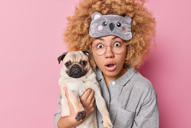 Stupefied scared young woman with curly blonde hair wears pajama and sleepmask holds pug dog reacts on shocking news isolated over pink background Emotional pet owner being afraid of something