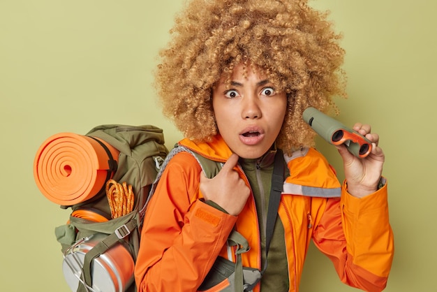 Stupefied shocked female explorer holds binoculars stares into distance carries backpack with equipment dressed in casual clothing isolated over green background People discovery exploration