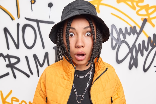 Ошеломленная девочка-подросток с дредами удивленно смотрит в камеру в черной шляпе, желтая куртка позирует на фоне разноцветной стены с граффити