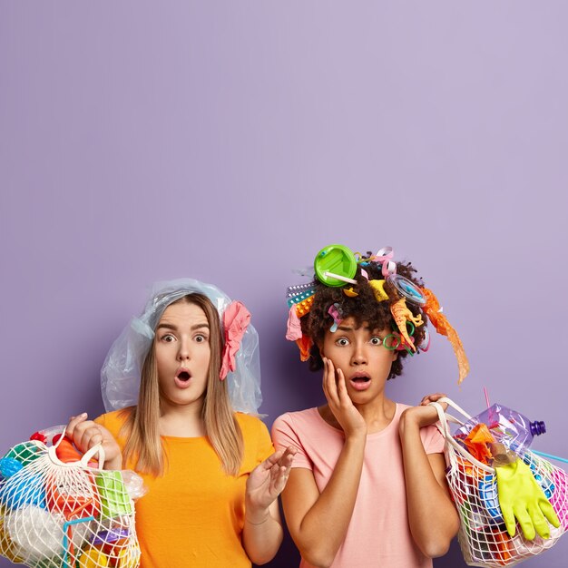 Stupefied two female activists stare with omg expression, shocked to pick up much garbage, hold net bags with plastic waste, dressed in casual outfit, pick up litter for recycling, free space above