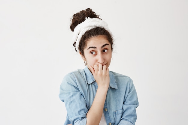 La donna stupida con scollo scuro e camicia di jeans ha un'espressione turbata, confusa o preoccupata, si morde le unghie, non vede scampo in una situazione difficile, si pone contro il muro bianco