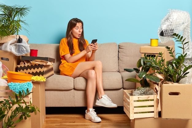 Stupefied young Caucasian houseowner moves to new apartment, changes home, stares at smartphone, makes plans for home improvement, sits on comfortable sofa, dressed in orange t shirt, white sneakers