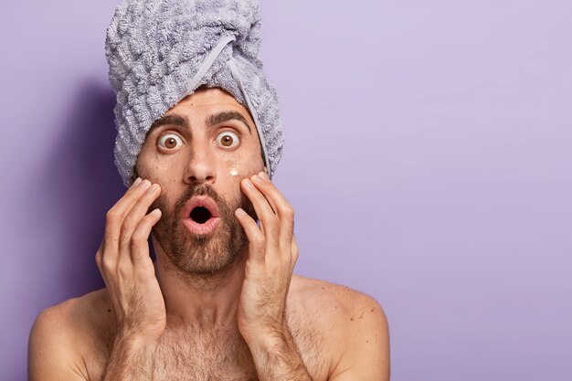 Stupefied young European man stares with bugged eyes, applies collagen pads under eyes, has bated breath