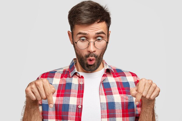 Stupefied young unshaven man wears casual shirt and eyeglasses, stares in surprisement down, points at floor