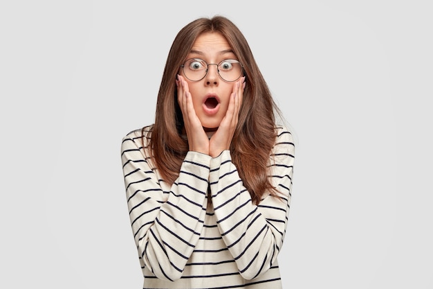 stupefied young woman with glasses posing against the white wall