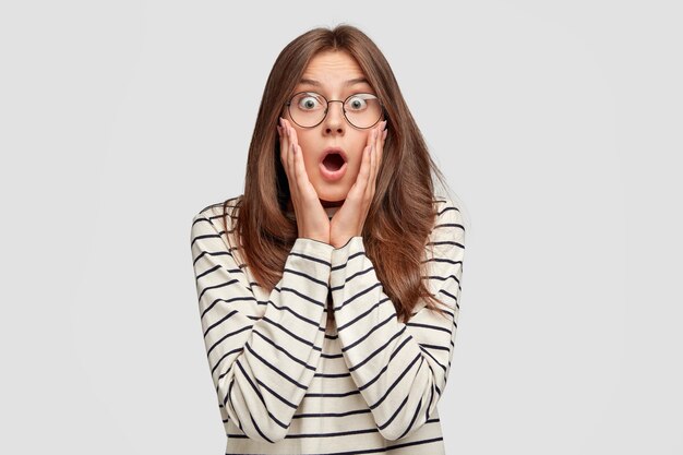 stupefied young woman with glasses posing against the white wall