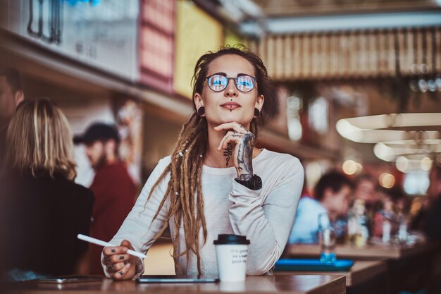 Стильная задумчивая девушка делает наброски в цифровом блокноте, сидя в кафе с чашкой кофе.