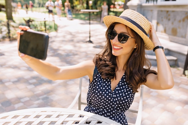 Style beautiful woman with short dark hair and charming smile is sitting in the summer cafeteria in sunlight. She is wearing summer hat and sunglasses and making selfie.