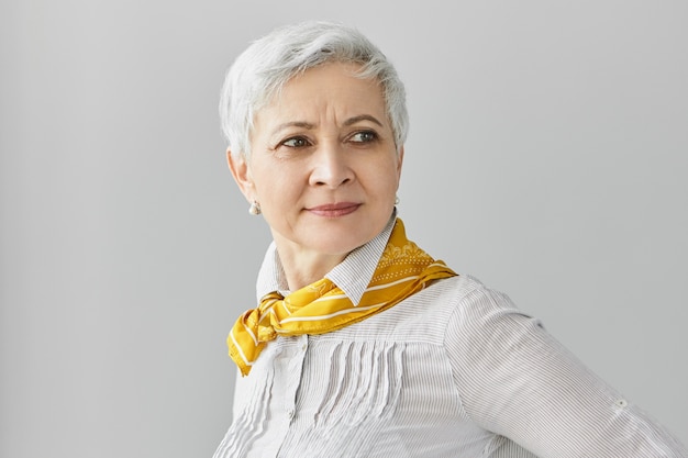 Free photo style, beauty and age concept. fashionable gray haired mature european lady wearing pearl earrings, blouse and yellow silk scarf around her neck posing, having confident facial expression