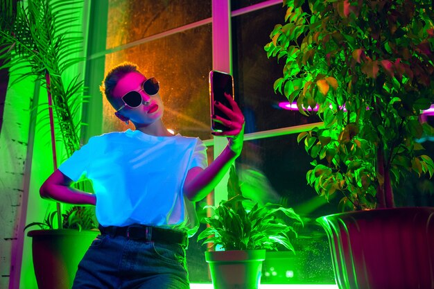 Style. Cinematic portrait of stylish woman in neon lighted interior. Toned like cinema effects, bright neoned colors. Caucasian model using smartphone in colorful lights indoors. Youth culture.