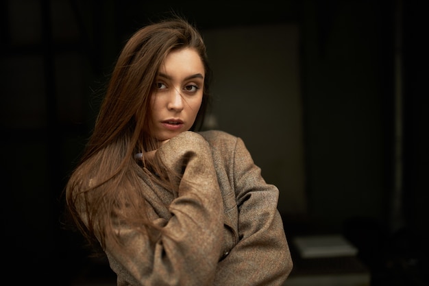 Style, fashion, beauty and femininity concept. Adorable gorgeous young woman with loose brown hair looking at camera with serious confident facial expression, dressed in oversize jacket or coat