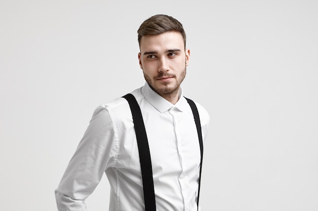 Style and fashion concept. Horizontal shot of attractive young European male model with mustache and stubble posing at white studio wall, dressed in elegant formal shirt with black suspenders