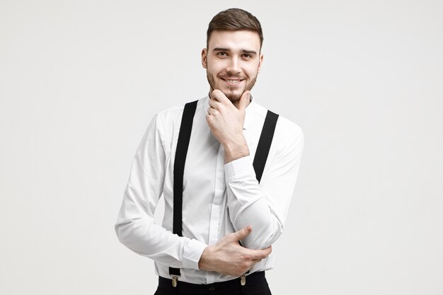 Style and fashion concept. Isolated picture of positive young unshaven male entrepreneur of Caucasian appearance wearing elegant formal clothes smiling cheerfully, touching his well trimmed beard