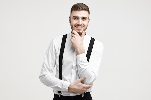 Концепция стиля и моды. Изолированная фотография позитивного молодого небритого мужчины-предпринимателя кавказской внешности в элегантной официальной одежде, весело улыбающегося, касаясь своей хорошо подстриженной бороды