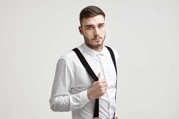 Style, fashion and elegance concept. Picture of fashionable young Caucasian bearded male manager wearing white shirt looking at camera with serious confident expression, pulling his suspenders