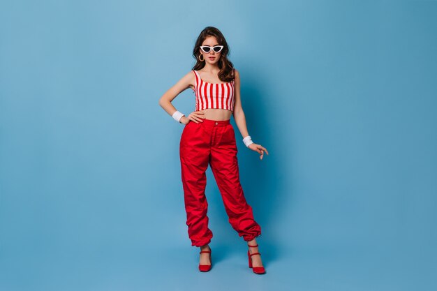 Stylish 80s woman in red pants and striped top poses on blue wall