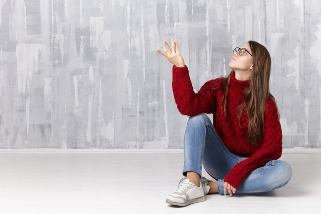 Stylish adorable long haired girl in warm knitted pullover, denim pants, sneakers and spectacles, gesturing while sitting comfortably on floor