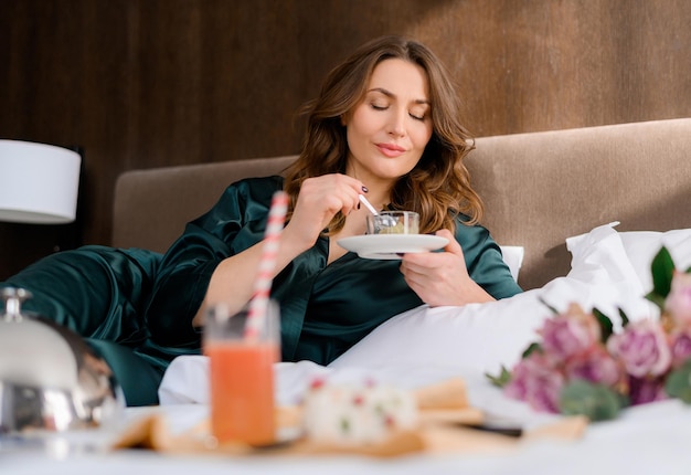 Стильная взрослая кавказская женщина ест десерт на кровати в комнате, имея свободное время Привлекательная дама носит зеленую пижаму, отдыхает и наслаждается праздниками Номер в отеле Красивая взрослая женщина