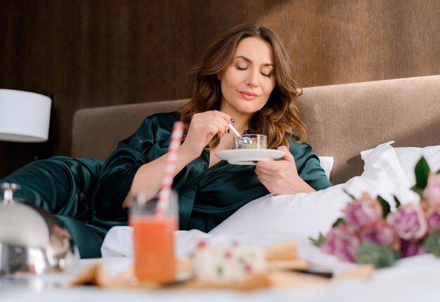 Stylish adult caucasian woman eating dessert on the bed in the room having free time Attractive lady wears green sleepwear resting and enjoying holidays Hotel room Beautiful adult woman